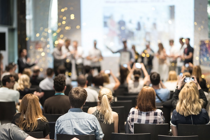 A group of people sitting in a class watching people on a stage.